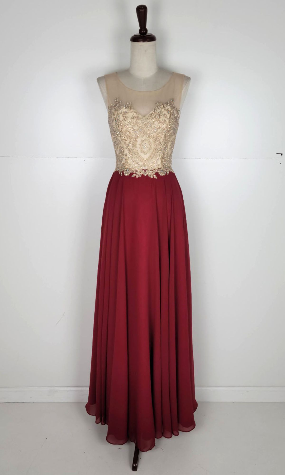 The Embroidered-Bodice Long Prom Dress: Chicas C3320 features a gold embroidered bodice, sheer neckline, and flowing burgundy A-line skirt, elegantly displayed on a dress form against a plain background.
